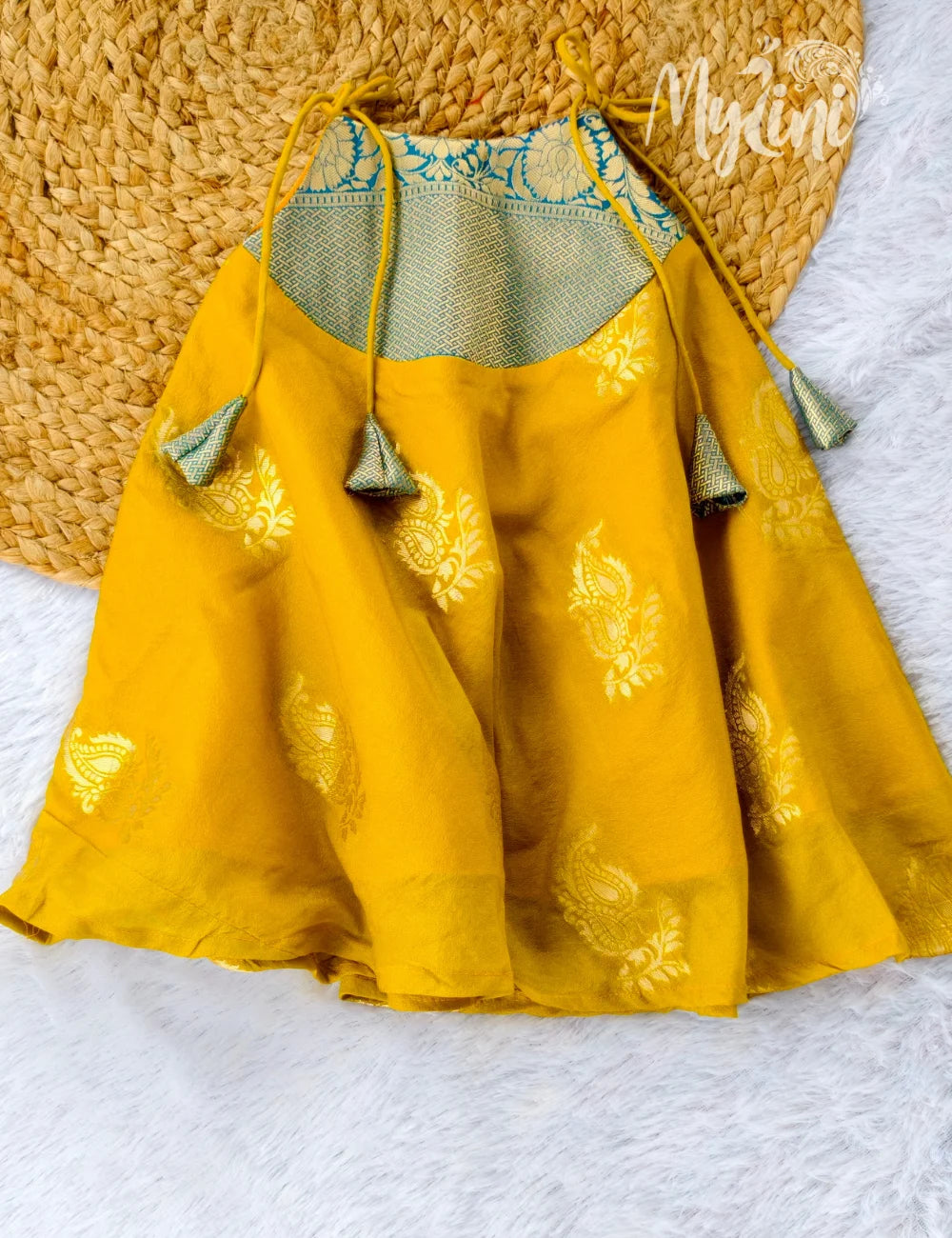 Baby in a green and yellow traditional outfit sitting on a white rug with a decorative background.
