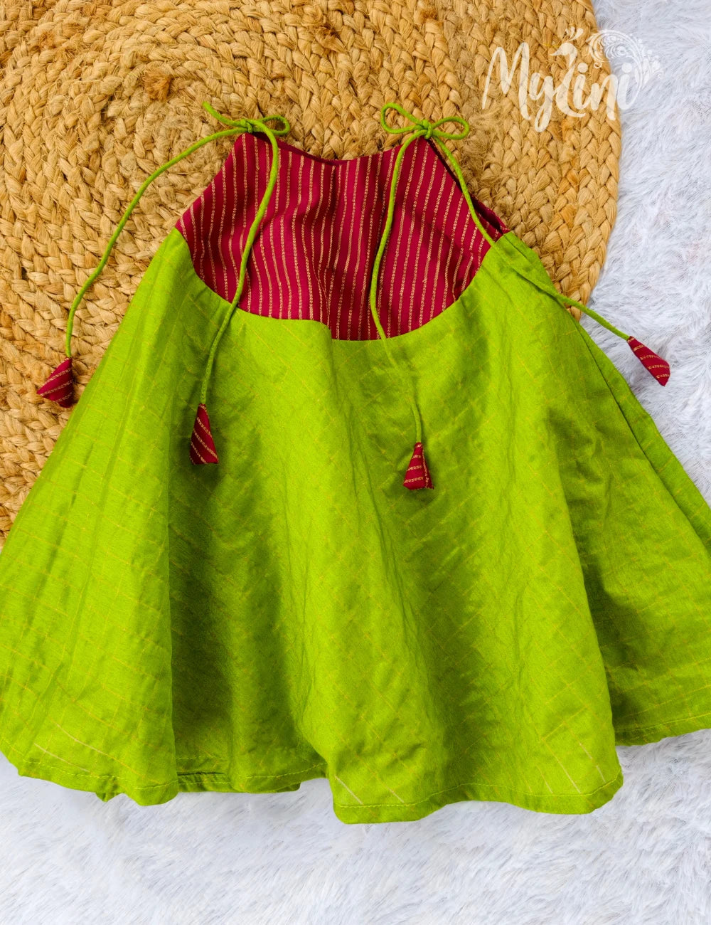 Baby wearing a red and green dress sitting on a white surface with a beige wall and decorative items in the background.