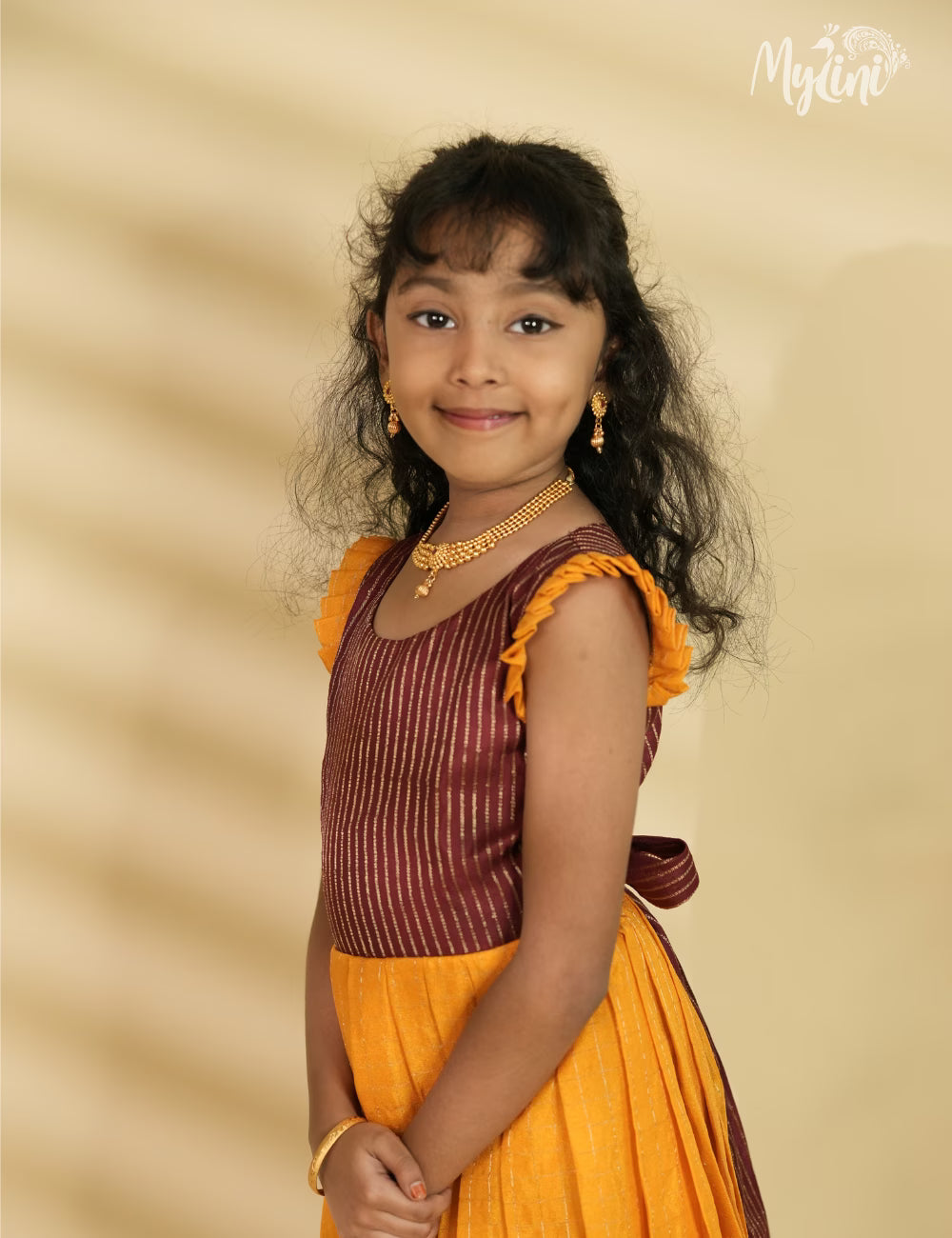 Young girl in a maroon and yellow traditional outfit sitting on a woven mat with a neutral background.