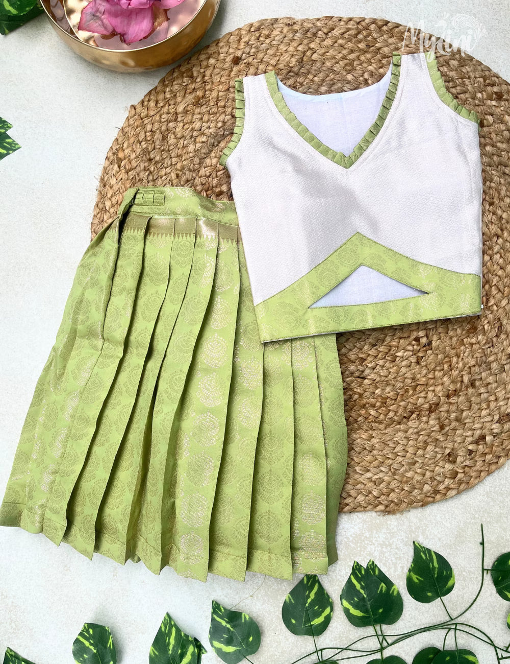 Young girl in a white and green traditional outfit standing on a woven mat with a beige background.