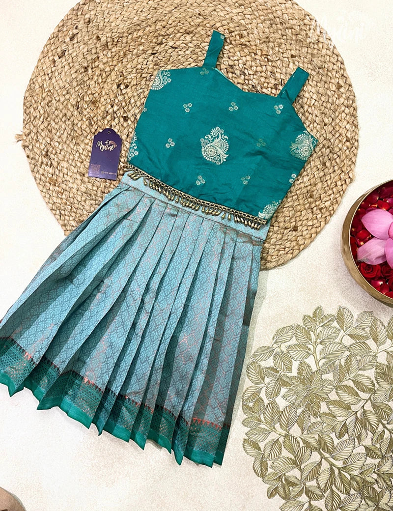 Child wearing a traditional outfit with a teal top and blue skirt in a decorated indoor setting.