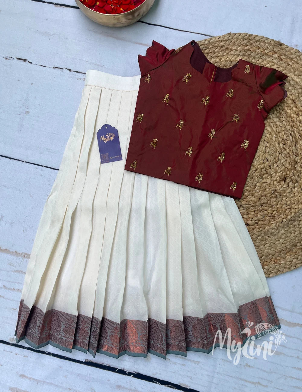 Young girl in a maroon and white traditional outfit standing on a mat with decorative items in the background.