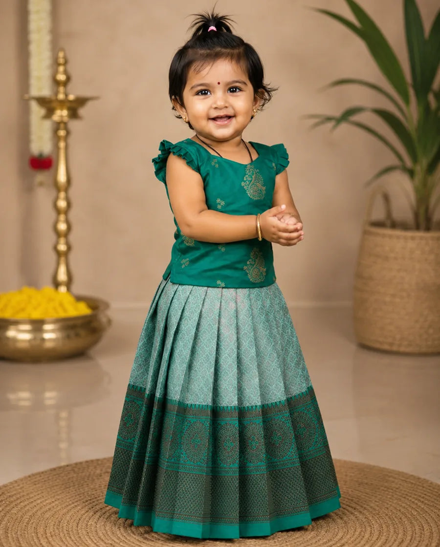 Young girl in a green traditional outfit standing indoors with decorative items in the background.