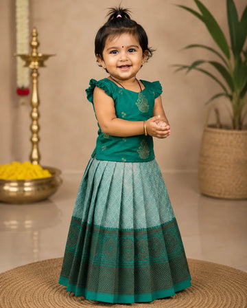 Young girl in a green traditional outfit standing indoors with decorative items in the background.