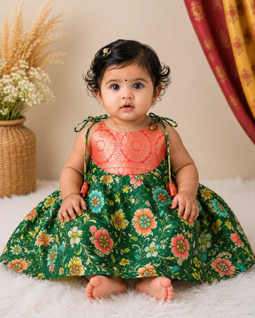 Baby wearing a floral dress with a pink top, sitting on a white surface with a neutral background.