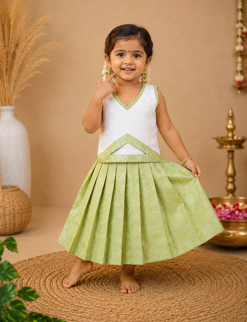 Young girl in a white and green traditional outfit standing on a woven mat with a beige background.