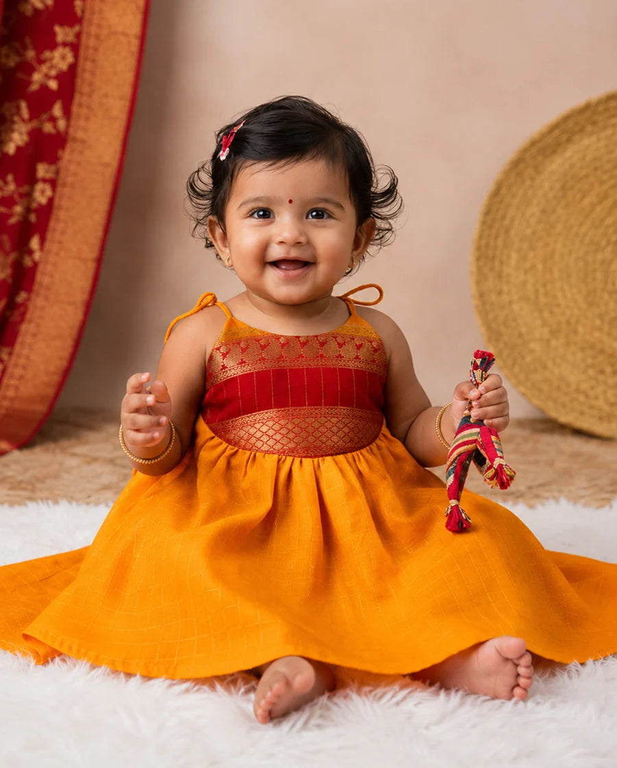 Baby in an orange dress with red accents sitting on a white surface.