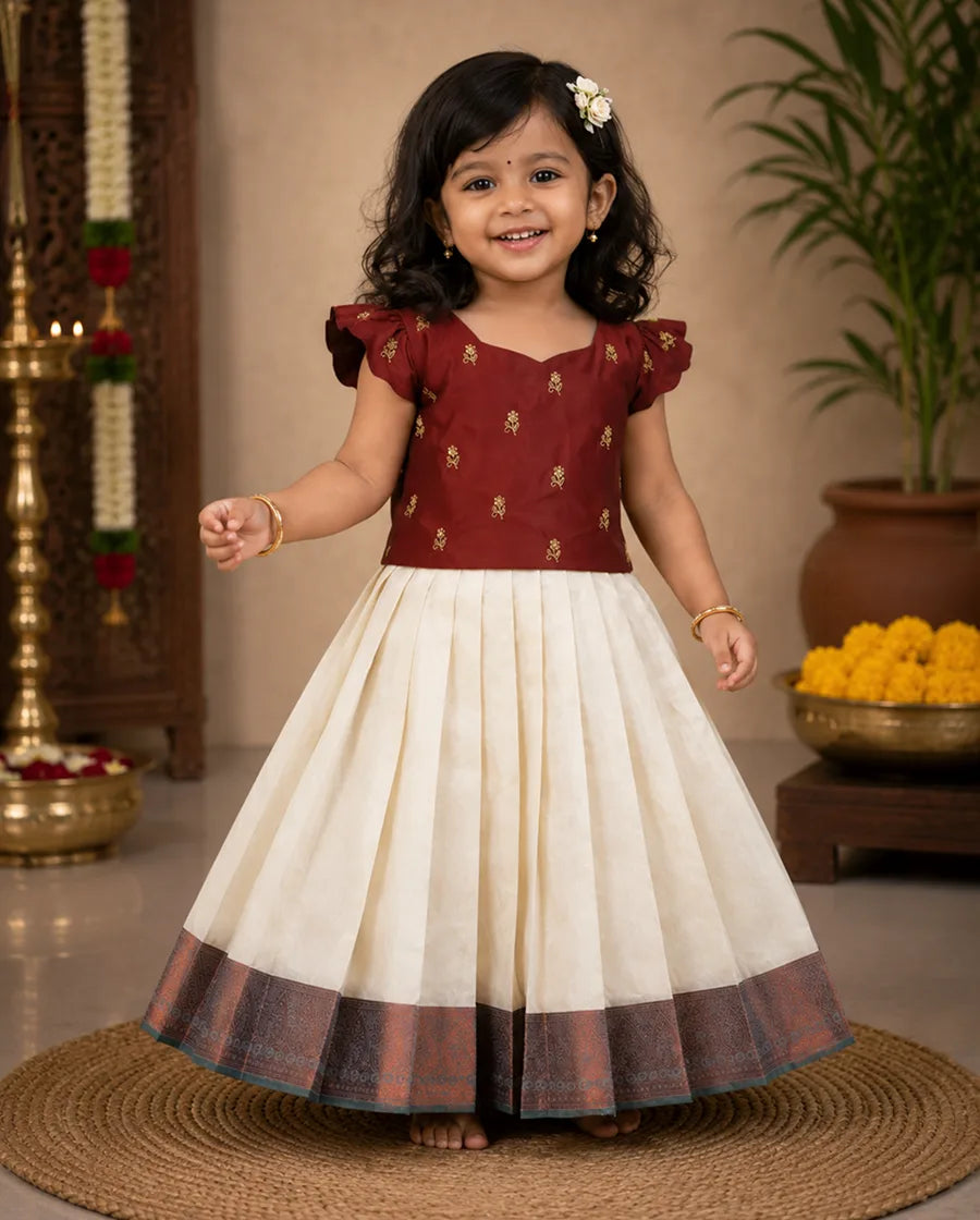 Young girl in a maroon and white traditional outfit standing on a mat with decorative items in the background.