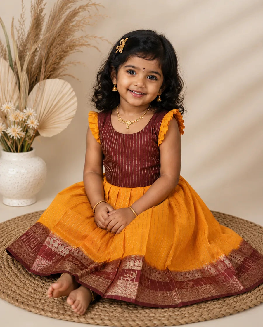 Young girl in a maroon and yellow traditional outfit sitting on a woven mat with a neutral background.