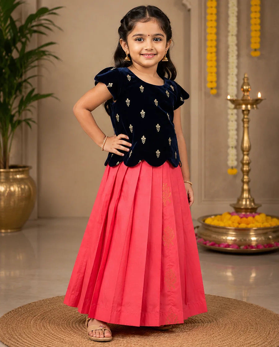 Young girl in a black top and pink skirt standing indoors with decorative elements in the background.