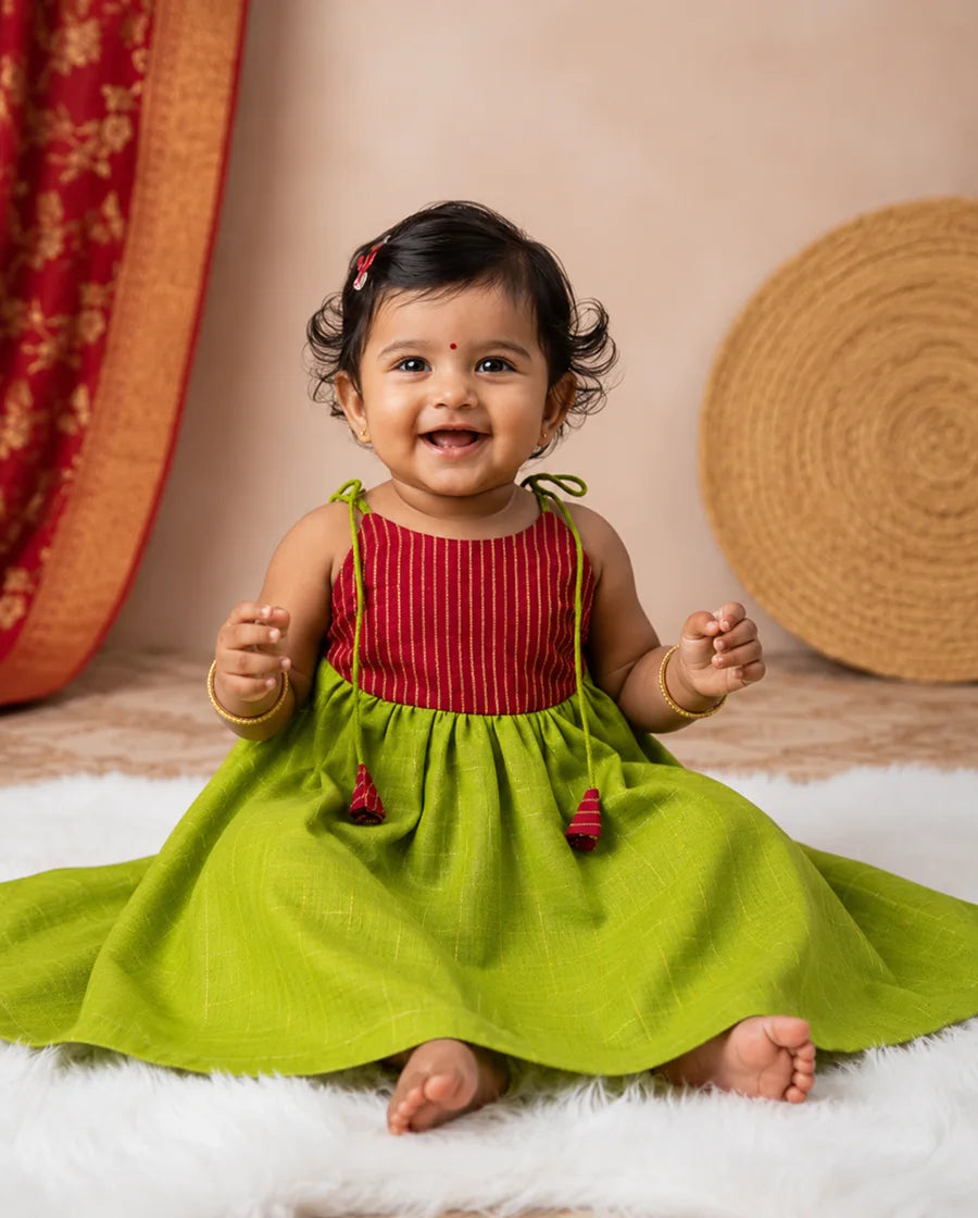 Baby wearing a red and green dress sitting on a white surface with a beige wall and decorative items in the background.