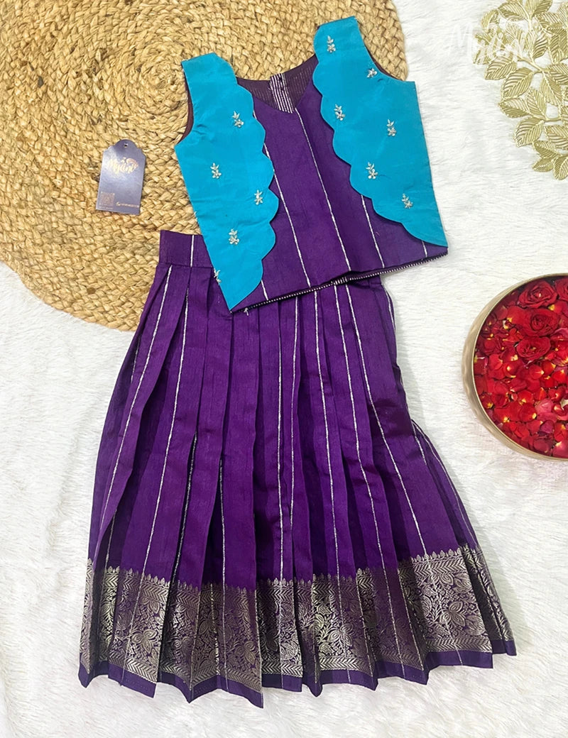 Young girl in a purple dress with a blue vest standing on a woven mat against a beige background.