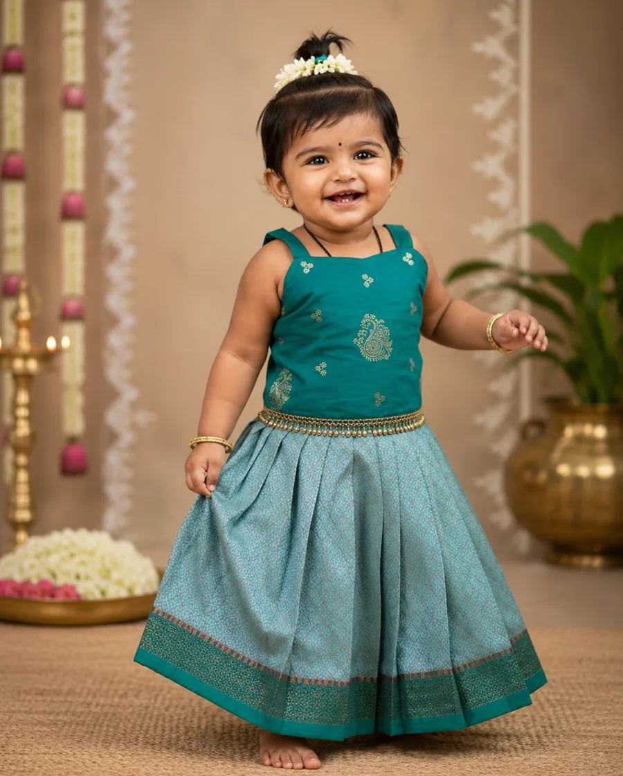 Child wearing a traditional outfit with a teal top and blue skirt in a decorated indoor setting.