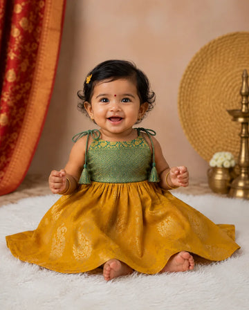 Baby in a green and yellow traditional outfit sitting on a white rug with a decorative background.